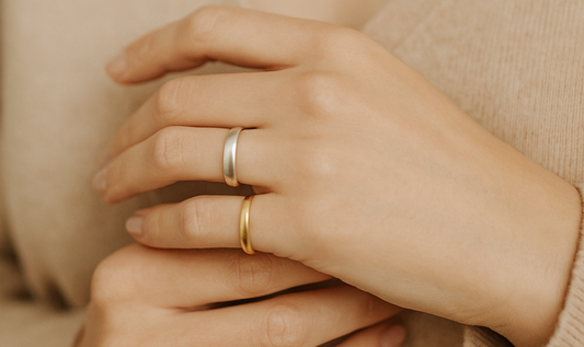 Main de femme portant deux bagues minimalistes, l'une en argent, l'autre en acier inoxydable, sur fond beige doux et naturel